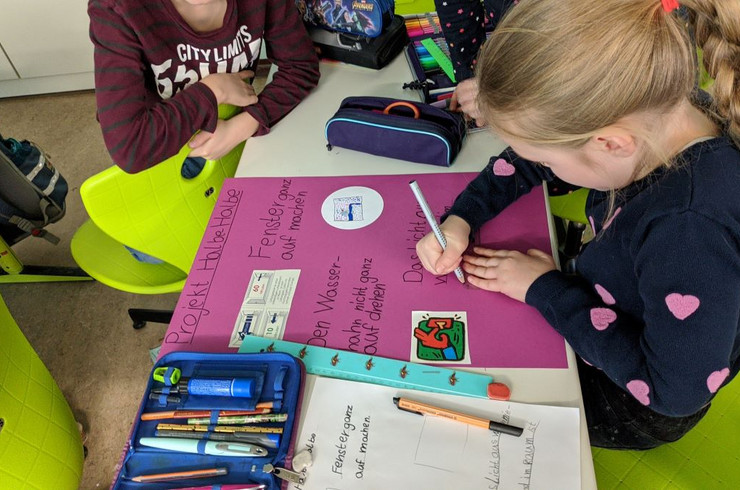 Ein Kind schreibt an einem lila Plakat, darauf sind Informationen und Bilder von Energiespartipps und die Überschrift "Projekt Halbe-Halbe"