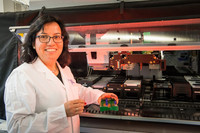 Fröhlich lachende junge Wissenschaftlerin mit dunklem Haar vor einer Apparatur