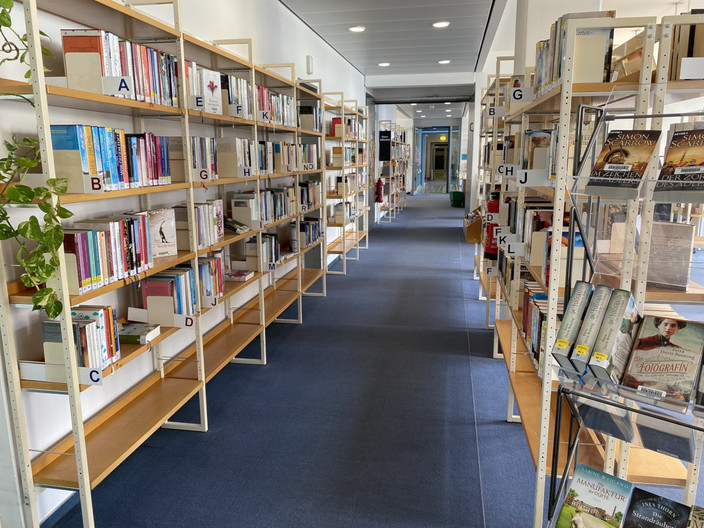 Ein Blick in die Bibliothek. Gang ist ausgelegt mit blauem Teppich. Links und rechts stehen viele Regale, samt Büchern. Hier ist die komplette Belletristik zu sehen. Mit Blick hinaus in den mit Glaskacheln verzierten Gang.