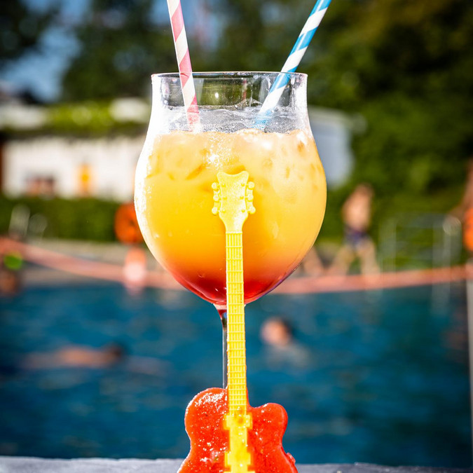 gelber Cocktail mit rotem Grund im Weinglas, zwei Strohhalme, aus Erdbeerpüree gefrorene Gitarre im Vordergrund