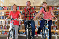 Drei Bibliotheksmitarbeiter auf ihren Fahrrädern stehen nebeneinander in der Bibliothek vor einem Bücherregal und lachen