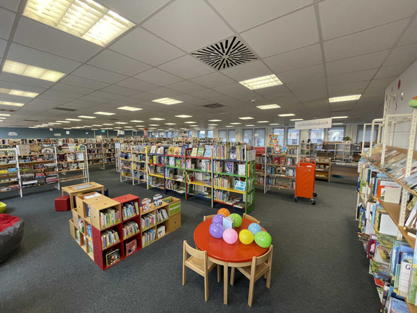 Weitläufiger Blick in die Bibliothek aus dem Kinderbereich. Im Vordergrund ein Kindertisch mit Stühlen und Luftballons.