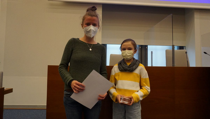 Eine Lehrerin mit Maske hält eine Urkunde in der Hand, rechts neben ihr eine Schülerin mit Maske und Schokolade in der Hand