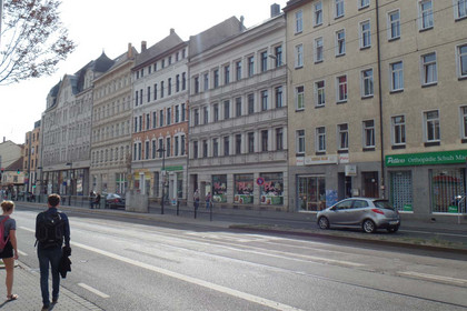 Die Georg-Schumann-Straße mit mehrstöckiger Häuserbebauung, Verkehrsanlagen der Straßenbahn, einige Passanten und Autos.