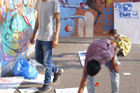 Jugendliche auf einem Hof mit Graffiti-bemalten Wänden. Ein Jugendlicher malt auf einem großen Blatt, das auf dem Boden liegt.