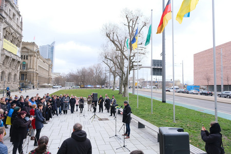 Links steht eine Gruppe Menschen vor einem großen alten Gebäude. Vor ihnen hält rechts im Bild ein Mann eine Rede. Hinter ihm sind einige Flaggen gehisst.