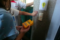 Kinder betrachten einen Lichtschalter, darüber ist ein Hinweisschild zum Licht sparen mit einer Glühbirne abgebildet, ein Kind hält ein Thermometer in der Hand