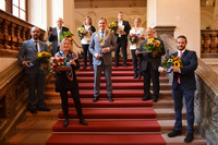 Eine Gruppe Menschen steht auf einer Treppe mit rotem Teppich, in der Mitte der Oberbürgermeister, die anderen halten Blumen und jeweils eine Ehrennadel in der Hand.