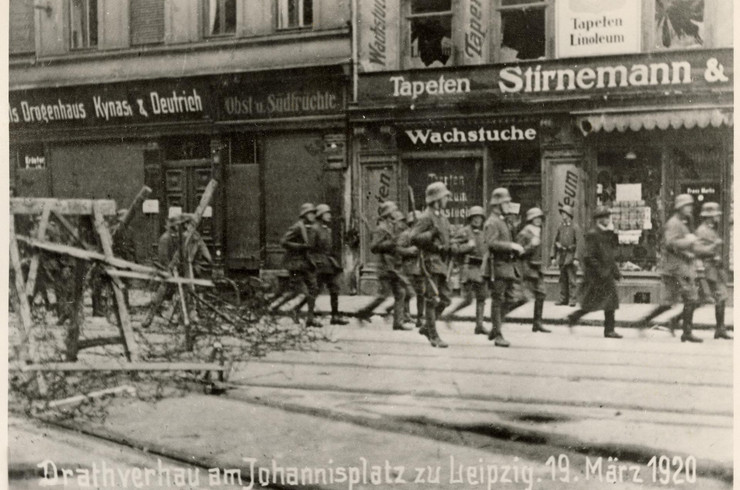 Das Bild ist eine historische Fotografie aus dem Jahr 1920. Es sind rennende Soldaten und ein Drahtverhau, der als Barrikade dient, auf der Straße am Johannisplatz in Leipzig zu sehen. Die Fotografie trägt das Datum 19. März 1920.