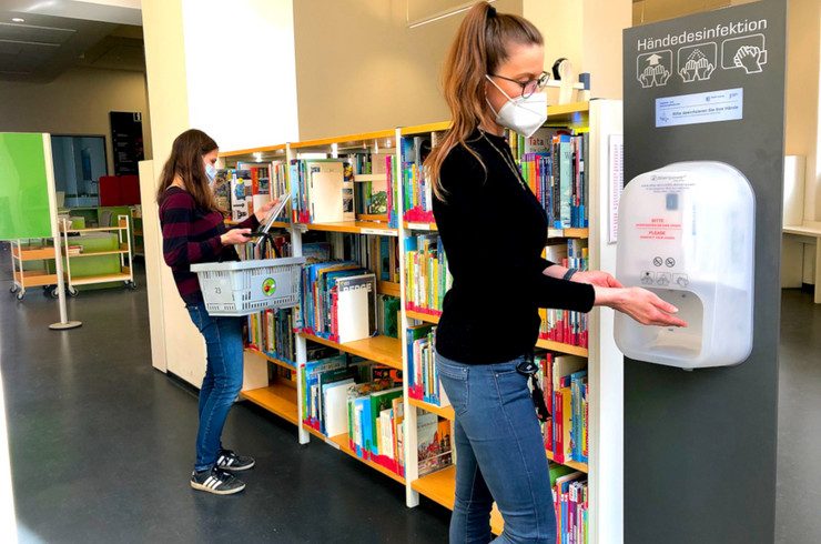 Innenraum der Leipziger Stadtbibliothek, eine Person benutzt einen Desinfektionsspender, eine zweite Person ist im Hintergrund bei der Suche nache einem passenden Buch, stehend an einem Regal zu sehen