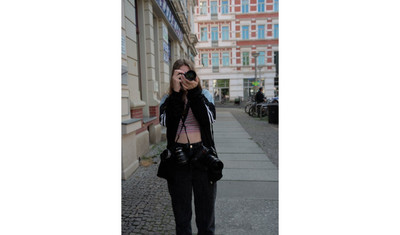 Eine Jugendliche in Alltagskleidung steht auf einem Gehweg in der Stadt und hält eine Spiegelreflexkamera in den Händen vor ihrem Gesicht. Sie hat zwei weitere Kameras umhängen.