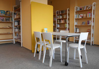 Ein weißer Tisch und weiße Stühle stehen in einem Bereich in der Bibliothek Grünau-Mitte. Im Hintergrund sind Regale mit Büchern zu sehen.