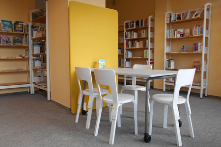 Ein weißer Tisch und weiße Stühle stehen in einem Bereich in der Bibliothek Grünau-Mitte. Im Hintergrund sind Regale mit Büchern zu sehen.