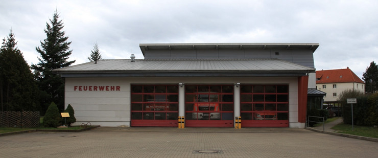 Eine Halle mit drei großen, roten Toren. In den Toren sind Fensterscheiben. Links neben den Toren eine weiße Wand, an deren oberem Ende in roten Buchstaben "Feuerwehr" steht.