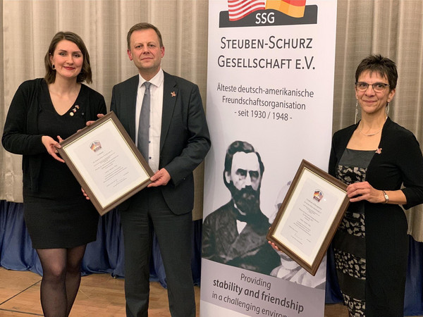 Herr Bonew und Frau Renner sowie Frau Langer halten zwei gerahmte Urkunden mit der Auszeichnung für die aktivste deutsch-amerikanische Städtepartnerschaft in 2018, daneben ein weißes Roll Up der Steuben-Schurz Gesellschaft