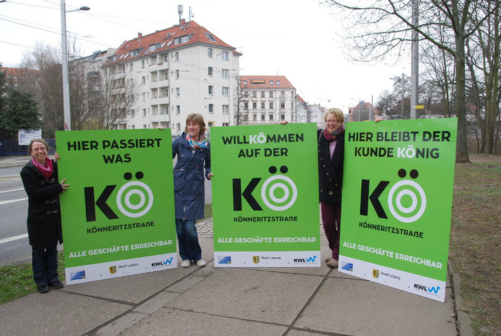 Drei Fraue halten die Hinweisschilder "Willkömmen auf der Kö" neben einer Straße