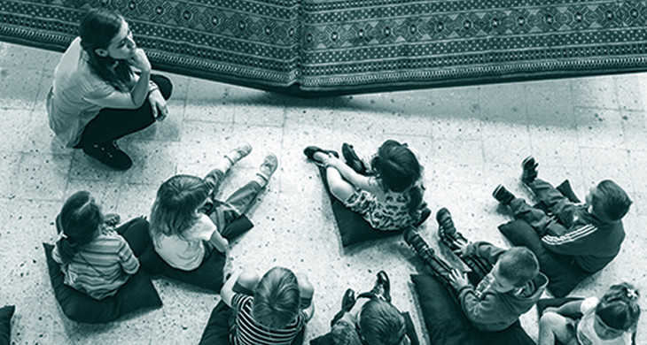 Acht Kinder sitzen auf Kisssen am Boden vor einem Kunstobjekt, eine Frau hockt davor und erklärt etwas.