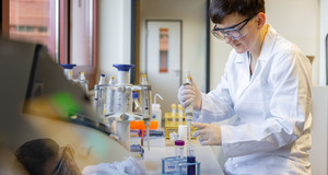 Eine Laborantin mit weißem Kittel und Schutzbrille sitzt an einem Tisch und entnimmt mit der Pipette eine Flüssigkeit aus einem Reagenzglas
