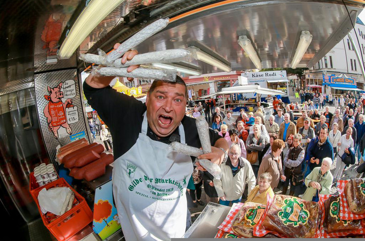 Marktschreier Wurst Achim hält Würste in der Hand und steht in seinem Verkaufswagen. Viele Menschen sehen ihm zu.