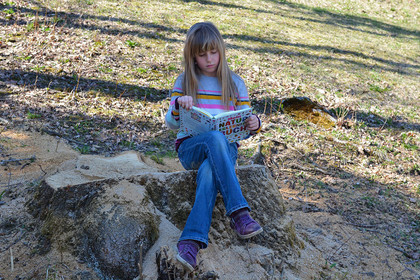 Ein Mädchen sitzt im Freien auf einem Baumstumpf und liest in einem Naturkundebuch.