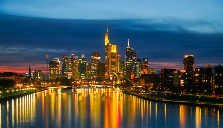 In später Abendstimmung sieht man die hell beleuchteten Hochhäuser Frankfurts im Main sich spiegeln