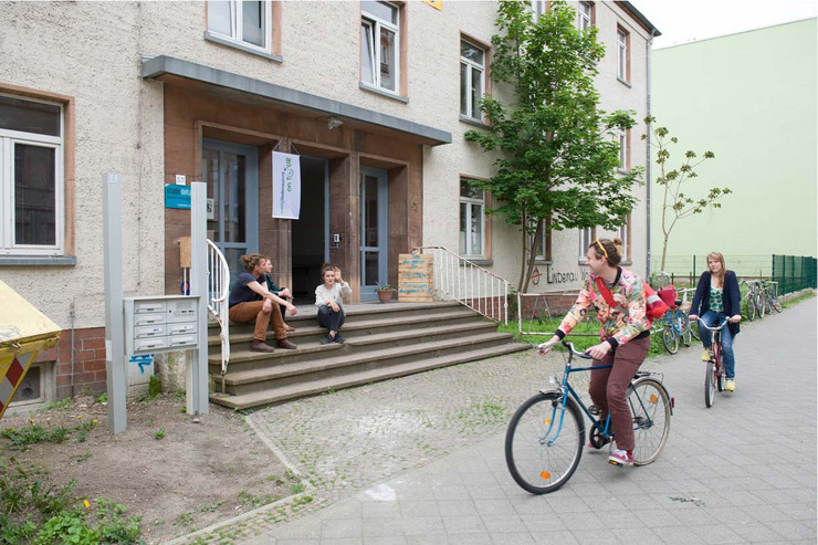 Zwei Fahrradfahrer fahren auf dem Fußweg vor einem Gebäude. Auf der Treppe vor dem Gebäude sitzen drei Menschen. Eine Person grüßt die Fahrradfahrer.