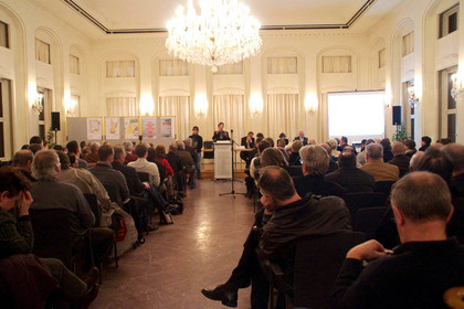 Zuhörer in einem Saal hören einem Redner zu, der die Ergebnisse der Jugendwerkstatt zum Leipziger Freiheits- und Einheitsdenkmal vorstellt.