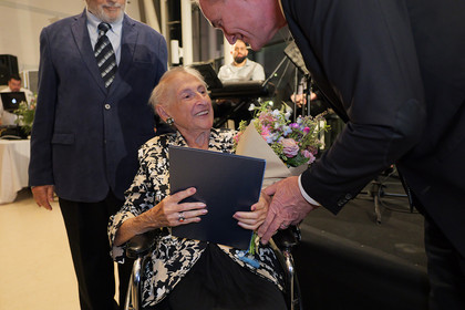 Eine ältere Frau im Rollstuhl erhält von einem Mann Blumen und ein Dokument überreicht.