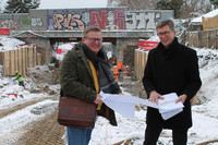 Zwei lachende Männer vor einer Baustelle halten gemeinsam einen Bauplan in den Händen.