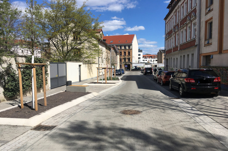 neugebaute Straße, links zwischen Fussweg und Straße befindet sich eine Pflanzinsel mit kleinen Bäumen, rechts ein Parkreihe mit Autos