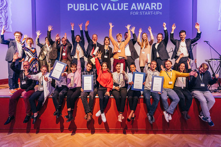 Das Foto zeigt die Gewinner des Public Value Award 2022 mit erhobenen Armen auf der Bühne