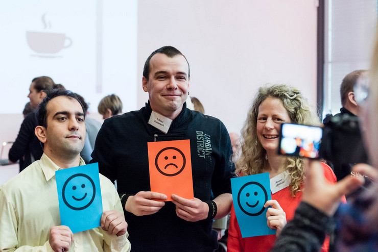 Zwei Männer und eine Frau lassen sich mit Karten fotografieren, auf denen Smileys abgebildet sind. Der Mann links und die Frau rechts halten eine blaue Karte mit lachendem Smiley hoch, der Mann in der Mitte eine orange Karte mit traurigem Smiley.