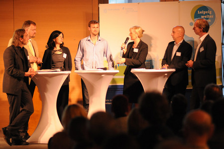 Podium während einer Veranstaltung steht an Stehtischen. Moderatorin hält Mikrofon in der Hand und redet mit den anderen Teilnehmern.