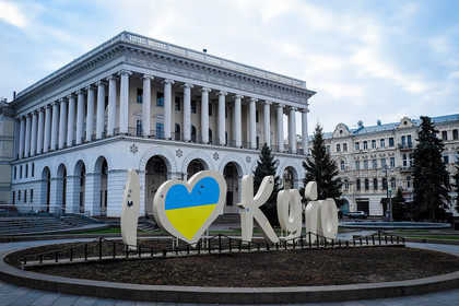 Der Schriftzug "I love Kyjiw", in dem das blau-gelbe Herz die Nationalfarben der Ukraine darstellt, vor einem weißen Gebäude mit Laubengang im Erdgeschoss und Säulen im oberen Gebäudeteil