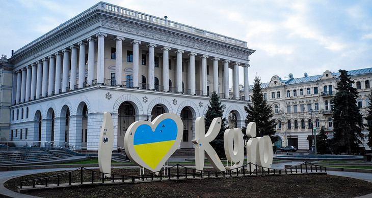 Der Schriftzug "I love Kyjiw", in dem das blau-gelbe Herz die Nationalfarben der Ukraine darstellt, vor einem weißen Gebäude mit Laubengang im Erdgeschoss und Säulen im oberen Gebäudeteil