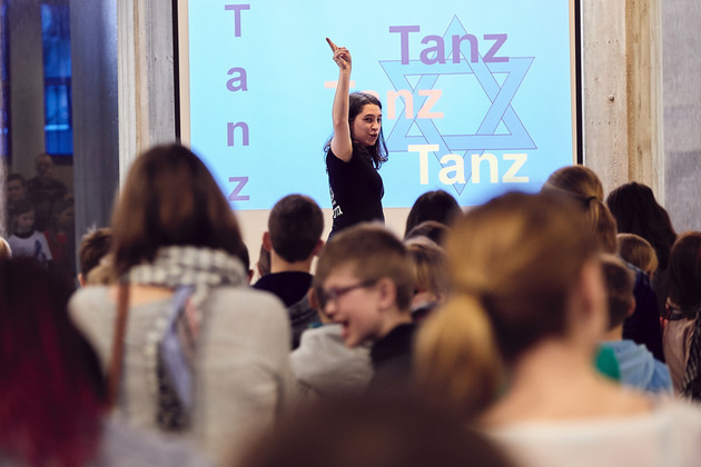 Vor einer Gruppe Jugendlicher steht eine junge Frau und reckt den rechten Zeigefinger am ausgetreckten Arm in die Luft, hinter ihr ein blaues Wandbild mit Davidstern und dem Schriftzug "Tanz"".