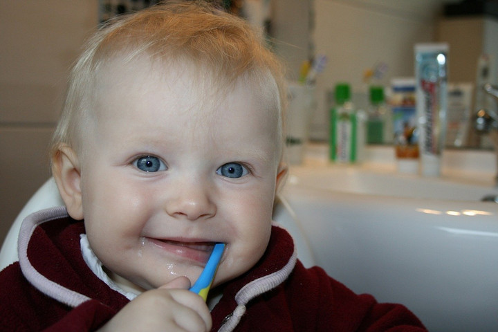 Kleines Kind mit einer Zahnbürste im Mund. Im Hintergrund ist ein Waschbecken.