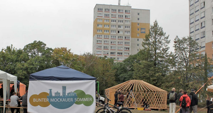 Blick auf das Gelände des Bunten Mockauer Sommers mit Pavillons und einer großen Plane mit dem Schriftzug "Bunter Mockauer Sommer". Im Hintergrund Hochhäuser