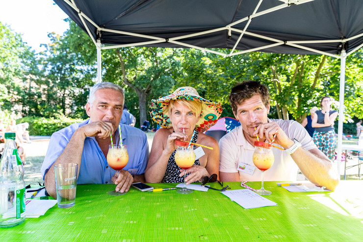 Zwei Männer und eine Frau mit Hut in der Mitte sitzen an einem Tisch, vor sich einen gelb-roten Cocktail, aus dem sie mit einem Strohhalm trinken.
