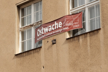 eine in die Jahre gekommene Fassade mit 2 Fenstern, zwischen den Fenstern befindet sich in Banner mit der Aufschrift: Ostwache