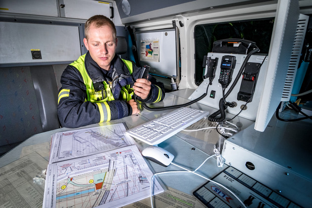 Ein Feuerwehrmann sitzt in einem Feuerwehrfahrzeug, vor einem Computer und einer Lagekarte, dabei hält er ein Funkgerät in der Hand