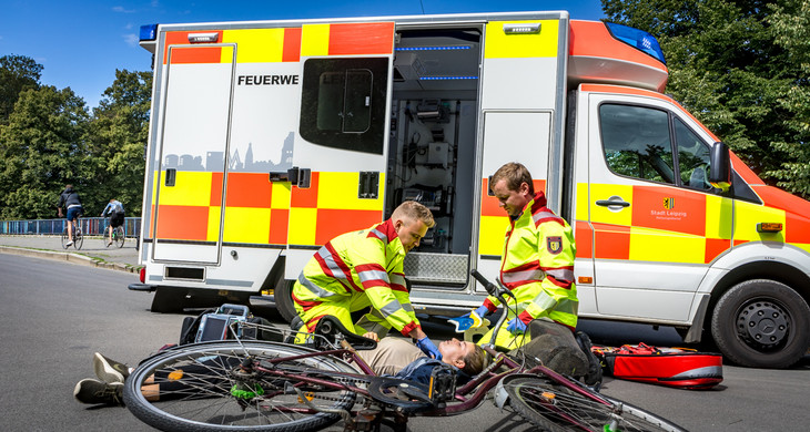 Zwei Notfallsanitäter versorgen eine gestürzte Radfahrerin. Die Radfahrerin liegt unter ihrem Fahrrad, die Rettungskräfte befinden sich neben ihr. Dahinter steht ein Rettungswagen mit geöffneter Seitentür.
