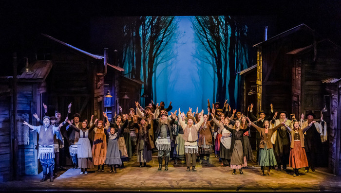 Eine Gruppe von Theaterdarsteller/-innen auf einer Bühne. Das Bühnenbild zeigt Holzhäuser und einen dunklen Wald.