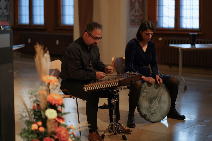 Musikgruppe SAMA Ensemble. Ein Mann mit einem Zupfgerät und eine Frau mit einer großen flachen Trommel