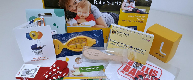 Das Leipziger Baby-Startpaket und sein Inhalt sind auf einem Tisch ausgebreitet. Mehrere kleine Geschenke wie zum Beispiel ein Babykalender, ein Badethermometer oder Babysöckchen sind zu sehen.