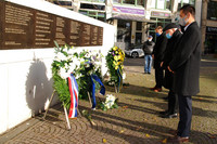 Fünf Männer stehen vor einer Mauer mit einer großen Gedenktafel, davor stehen drei große Blumenkränze.