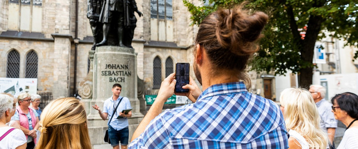 Eine Gruppe Touristen steht vor dem Bach-Denkmal im Thomaskirchhof, ein junger Mann macht Fotos von dem Denkmal, vor dem ein Gästeführer steht.