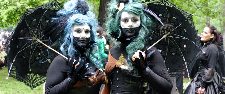 Zwei Frauen mit schwarzen Kleidern, schwarzen Spitzenschirmen, weiß geschminkten Gesichtern und blauen beziehungsweise grünen Haaren