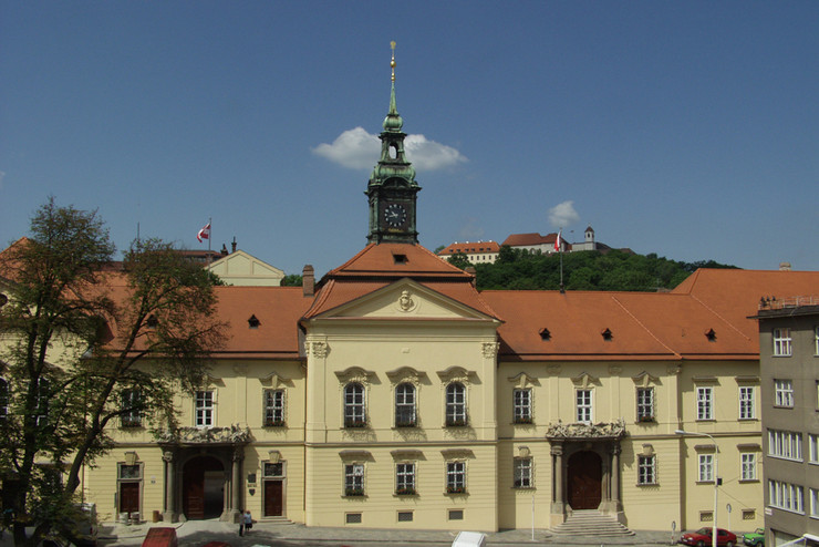 gelbes schlossähnliches Gebäude mit rotem Dach vor blauem Himmel und Burg Spilberk im Hintergrund