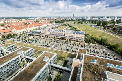 Blick von oben auf das MDR-Gelände und MediaCity mit vielen Gebäuden, einem Parkplatz und weit dahinter der Leipziger Innenstadt